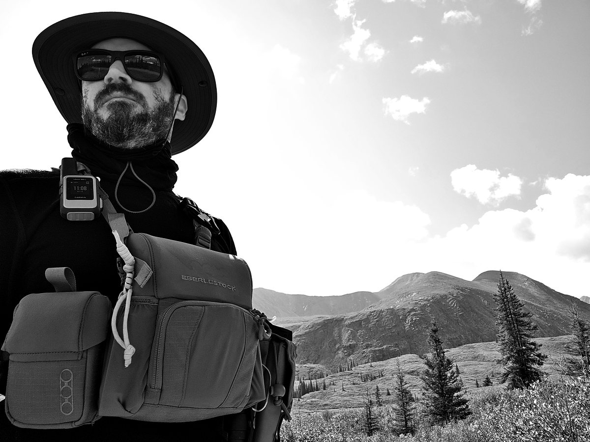 André in a wide-brimmed hat and a chest pack, mountains behind him
